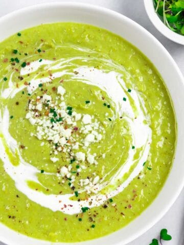 White bowl of creamy broccoli almond soup topped with a swirl of sour cream, and sprinkled with crumbly cheese and herbs.