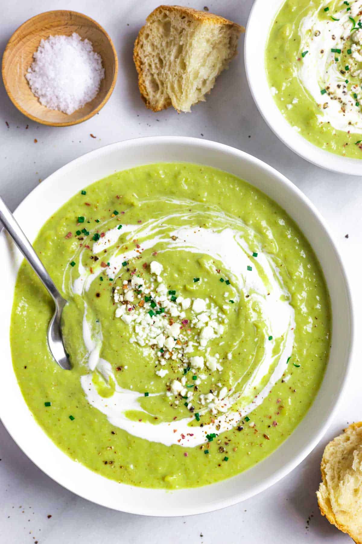 White bowl of broccoli almond soup with a spoon, sour cream, cheese and herb topping.