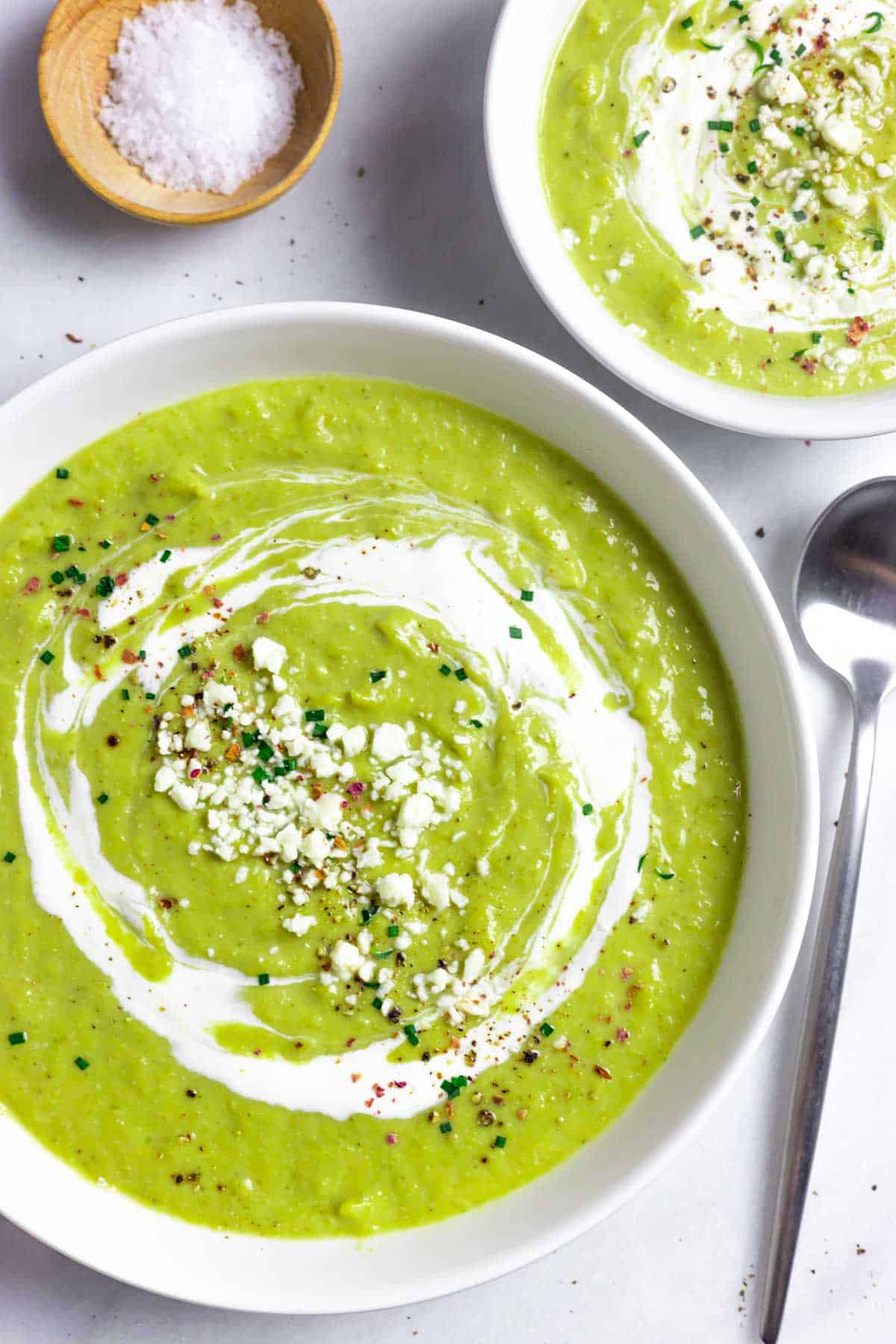 Two bowls of blended broccoli almond soup topped with sour cream, cheese and herbs.