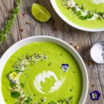 Two bowls of avocado gazpacho decorated with sour cream, sprinkled herbs and edible flowers.