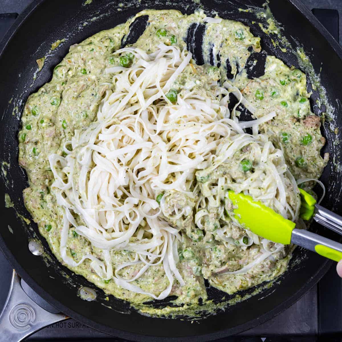 Folding noodles into pesto sauce in a skillet with tongs.