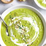 White bowl of broccoli almond soup with a spoon, sour cream, cheese and herb topping.