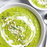 Two bowls of blended broccoli almond soup topped with sour cream, cheese and herbs.