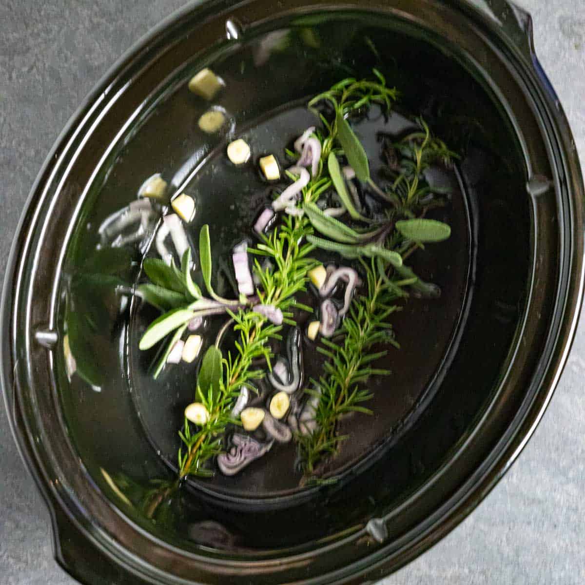 Fresh herbs in bottom of a crock pot including sage, rosemary and shallots.