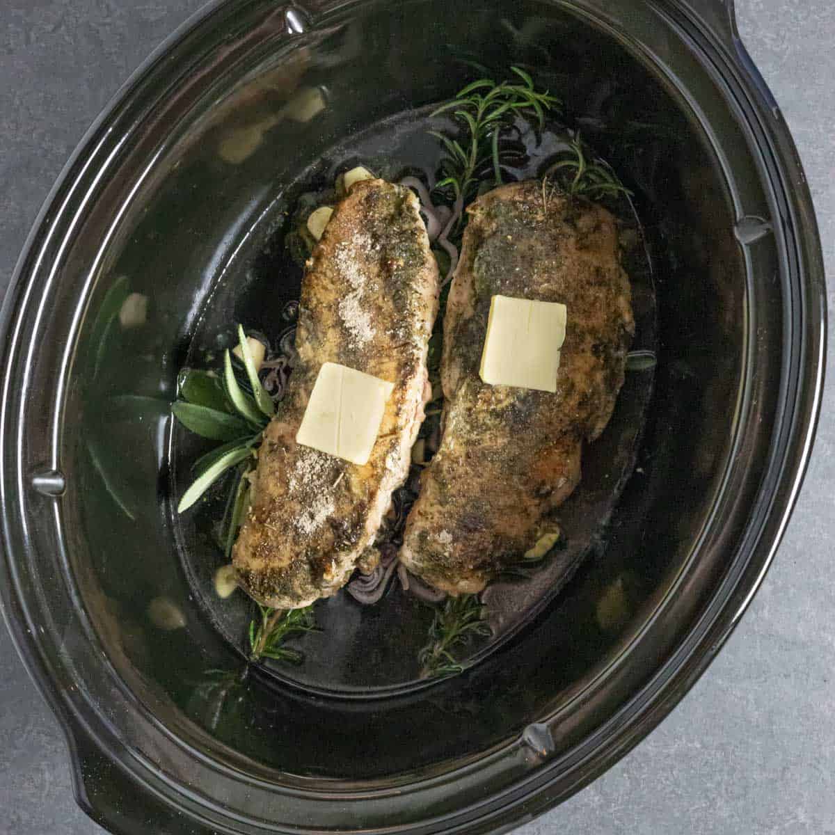 Turkey breast tenders with a pat of butter on top in a slow cooker over fresh herbs.