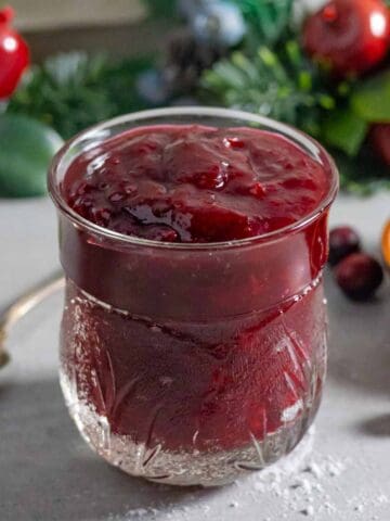 Fancy jar of bright red cranberry jam with festive greens behind.