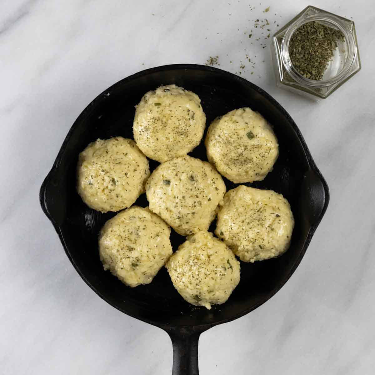 Balls of dough in a small cast iron pan topped with dried parsley and ready to bake.