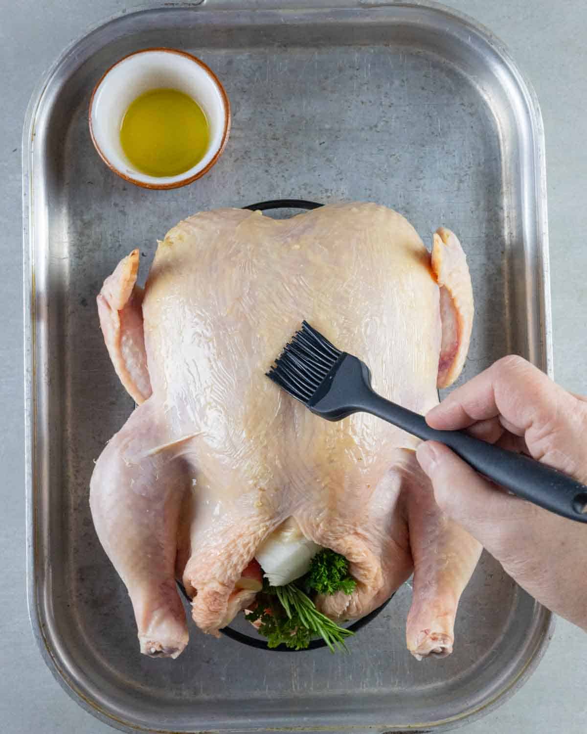Whole chicken in a roasting pan being brushed with melted butter.