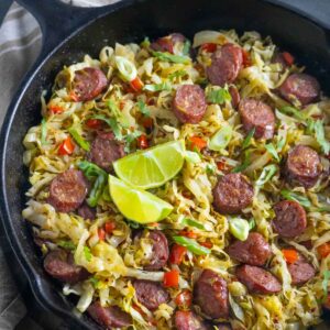 Fried cabbage and sausage skillet garnished with lime wedges.