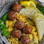 Plate of corned beef meatballs on top of colcannon with a pan-fried wedge of cabbage, lemon wedges and parsley.