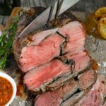 Boneless leg of lamb being sliced on a board with grilled lemon and a small bowl of red chimichurri sauce.
