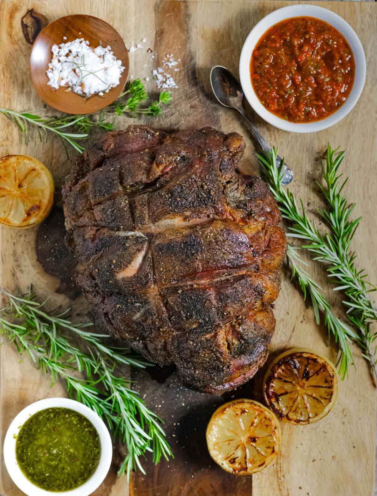 Whole roast lamb leg with crusty bark on a board with rosemary sprigs, ramekins of chimichurri sauce and grilled lemons.