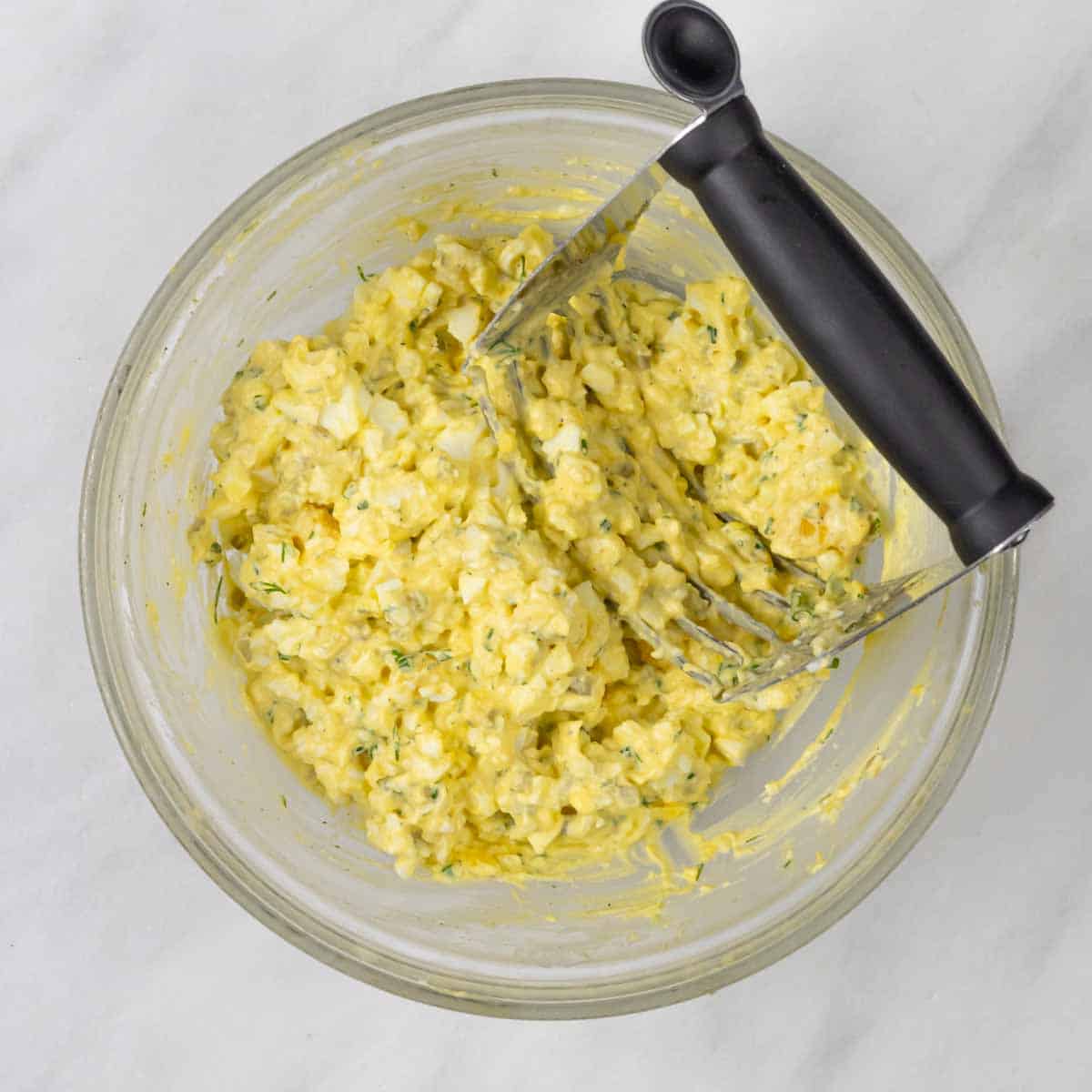 Ingredients for egg salad mixed in a glass bowl using a pastry cutter.