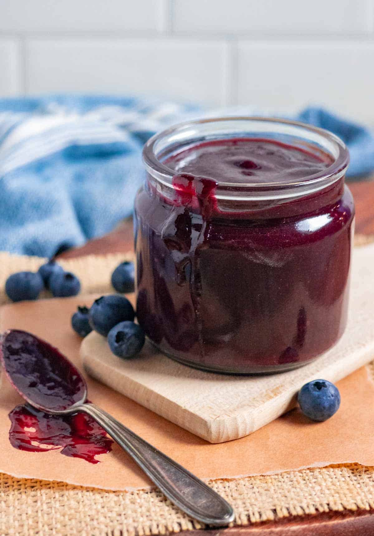 Glass jar of blueberry bbq sauce with dribble down its side on a board with blueberries and a sauced up spoon.