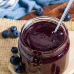 Blueberry bbq sauce in a jar with a spoon sticking out, on a board with fresh blueberries scattered around.