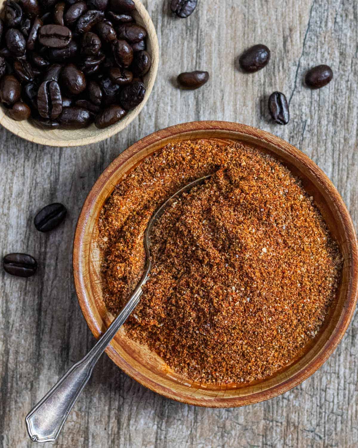 Coffee rub in small wood bowl with a spoon in it next to a small bowl of whole coffee beans.