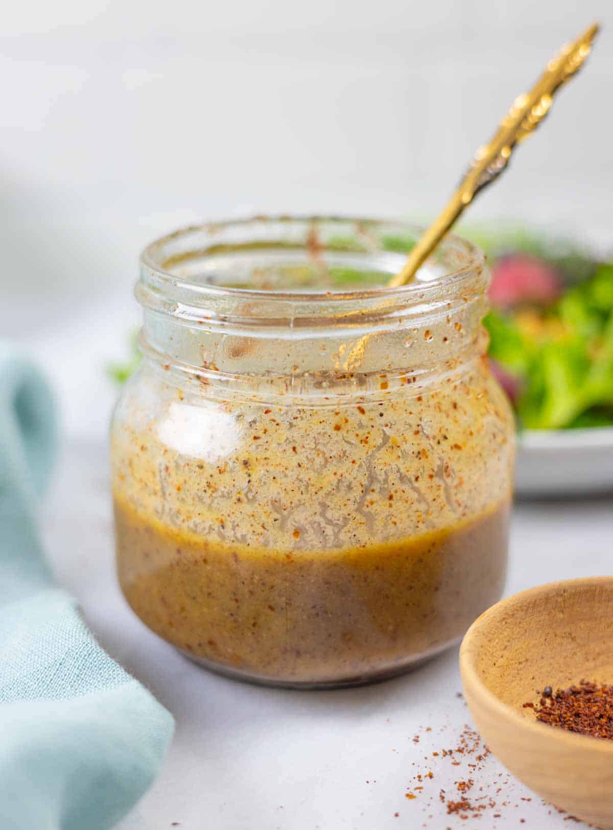 Jar of sumac dressing with a spoon in it and bowl of salad behind.
