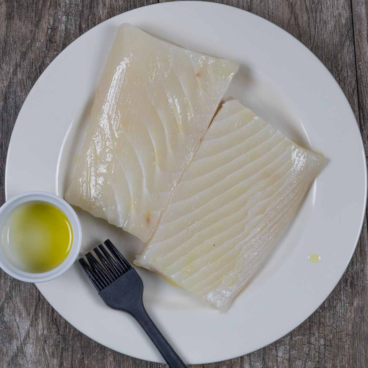 Two halibut filets on a white plate with a ramekin of oil and basting brush.