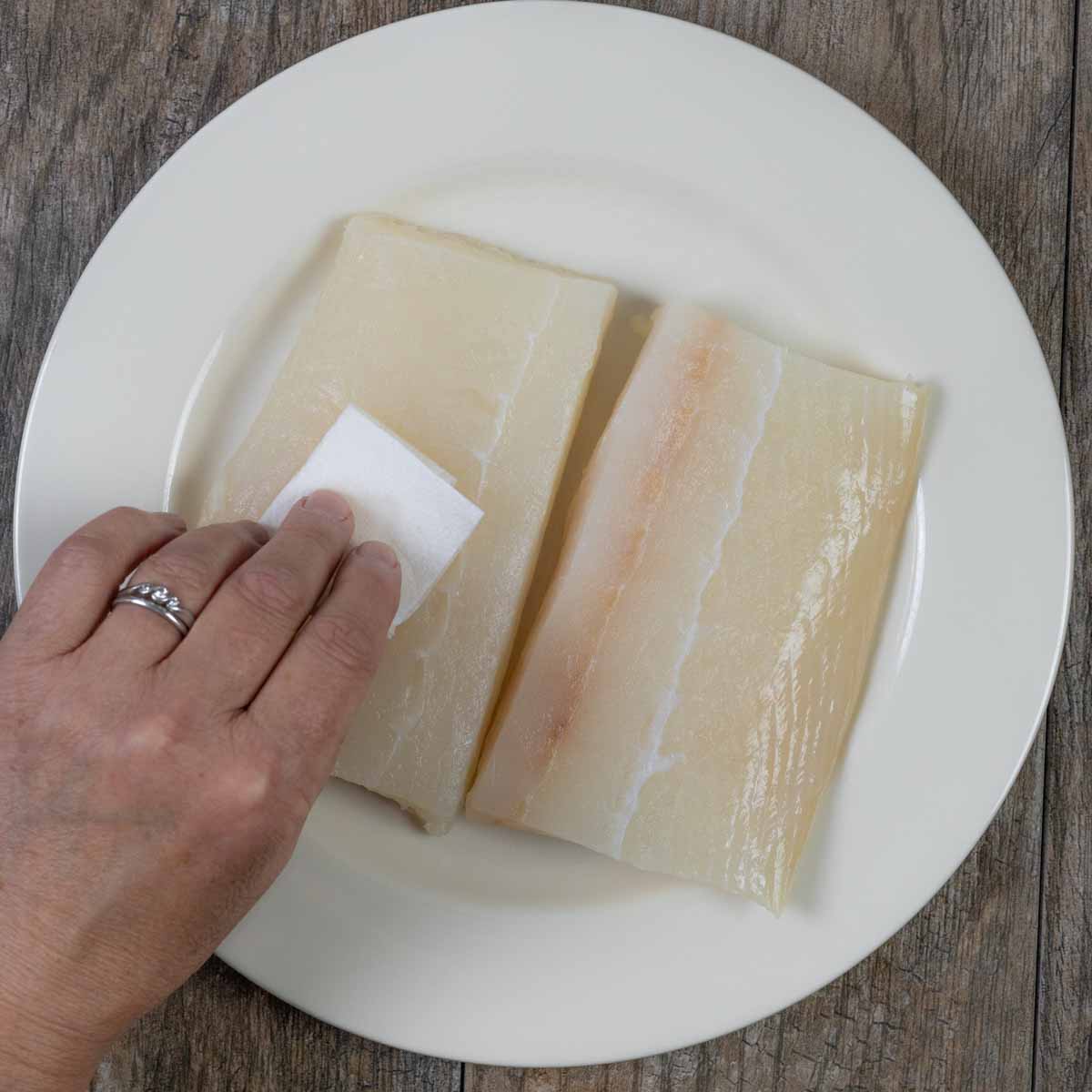 Filets of halibut on a white plate with a person's hand patting dry with a paper towel.