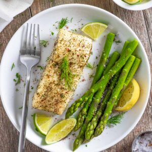 Air fried halibut with herbs and citrus on a white plate with asparagus spears and a fork.