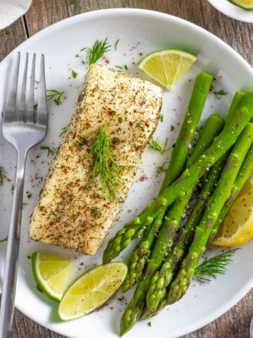 Air fried halibut with herbs and citrus on a white plate with asparagus spears and a fork.