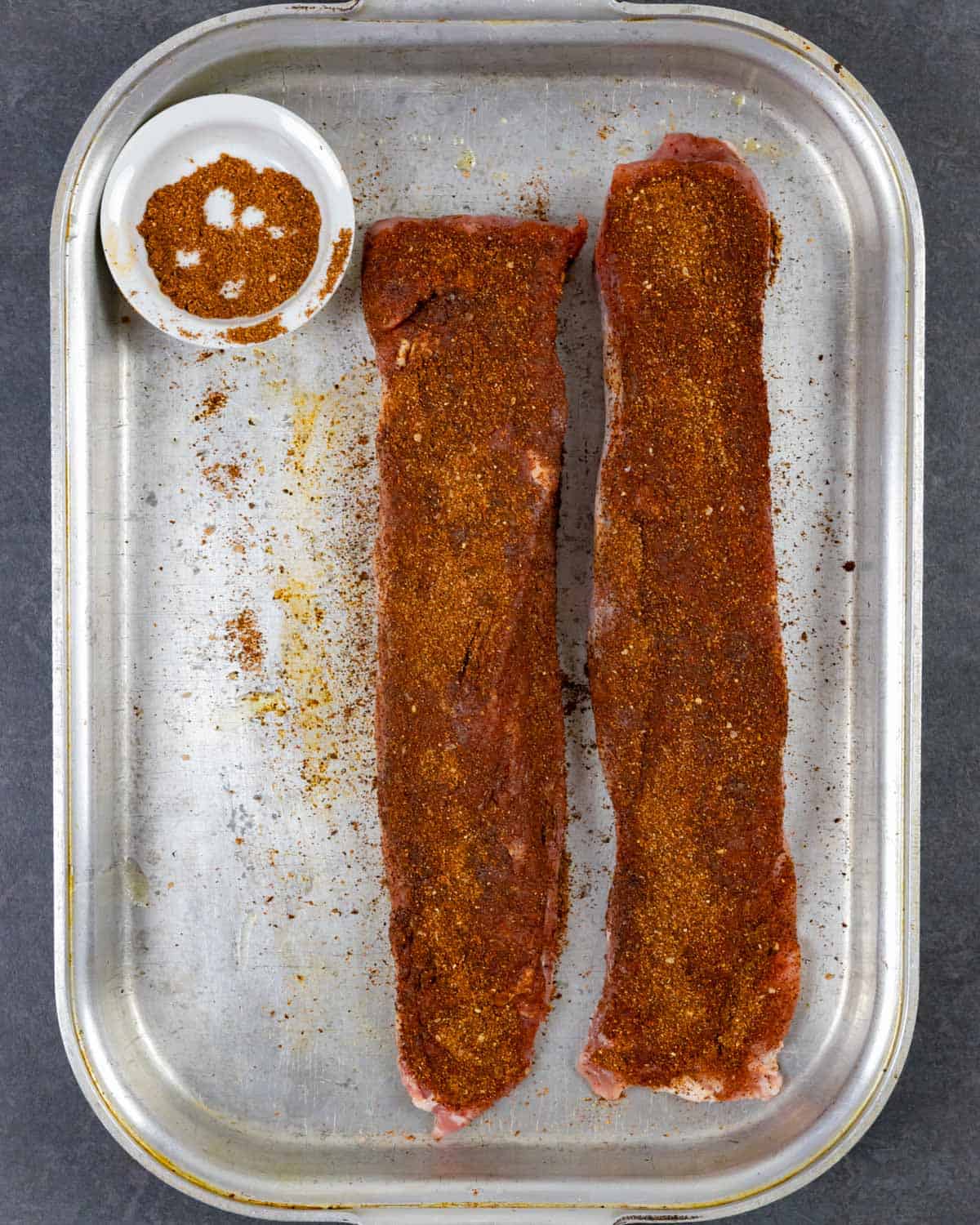Two pork tenderloins in a metal pan coated with dry rub next to a ramekin with rub in it.