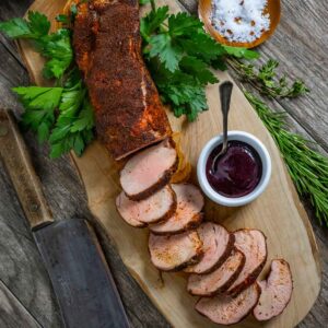 Smoked pork tenderloin sliced on a board with ramekin of blueberry barbecue sauce.