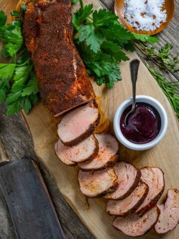 Smoked pork tenderloin sliced on a board with ramekin of blueberry barbecue sauce.