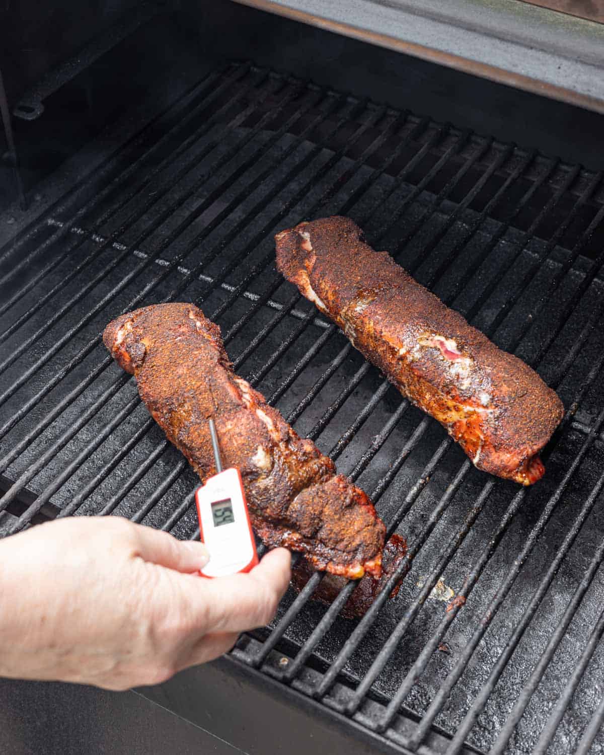 Two pork tenderloins on the smoker grates with thermometer inserted checking internal temperature.