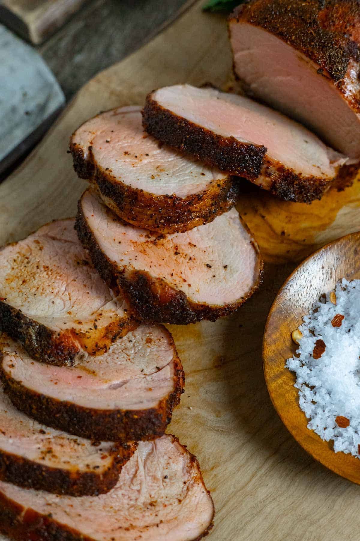 Slices of smoked pork tenderloin on a board next to a wood bowl of flake salt.