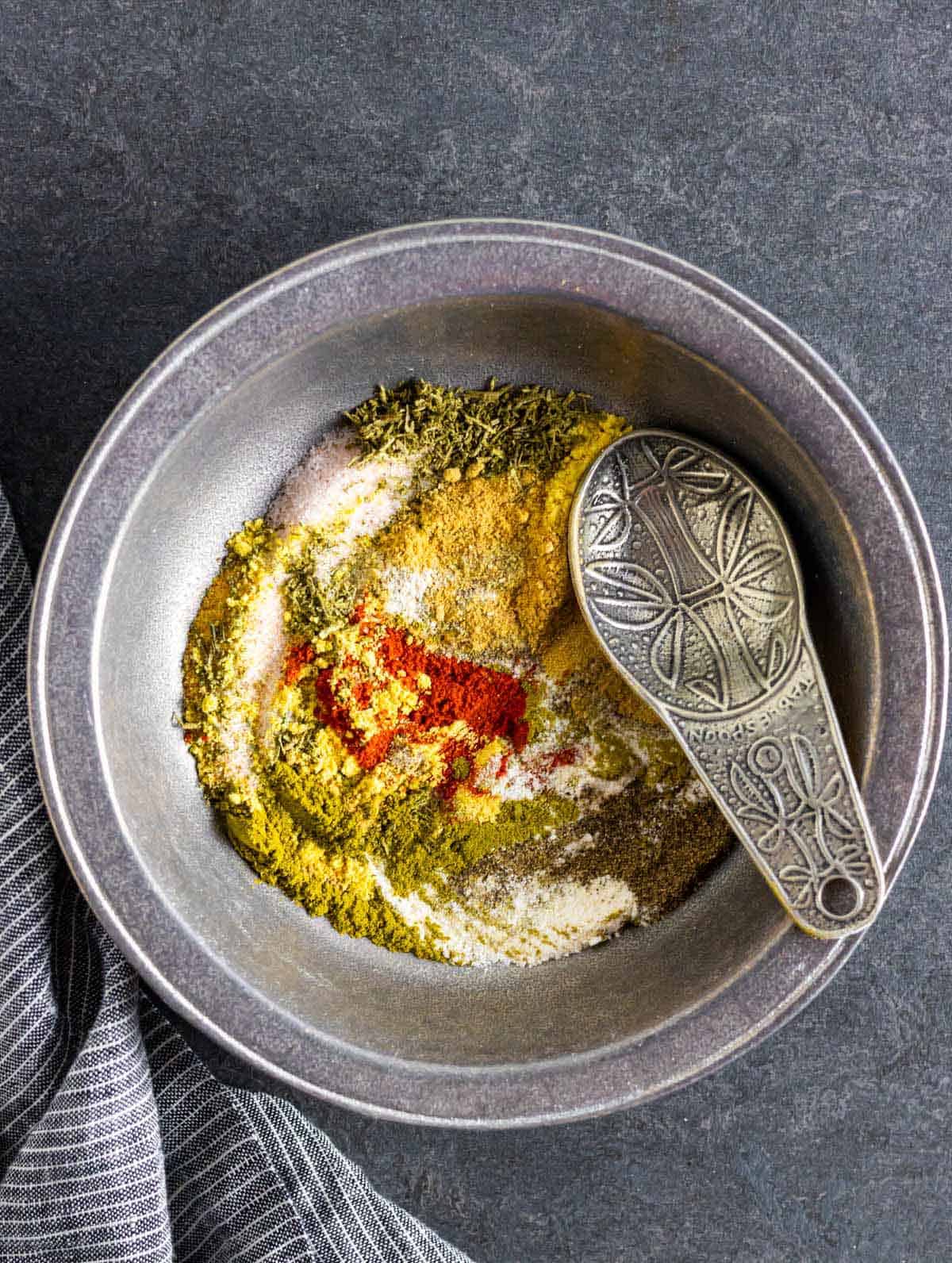 Homemade beef spice rub in a pewter bowl partially stirred with a fancy pewter measuring spoon.