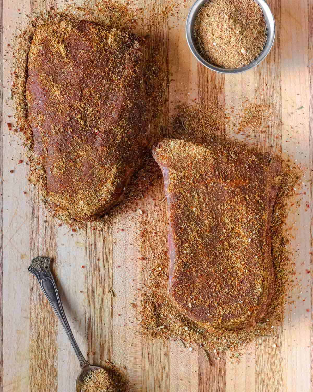 Two raw sirloin steaks on a board covered in beef dry rub seasoning.