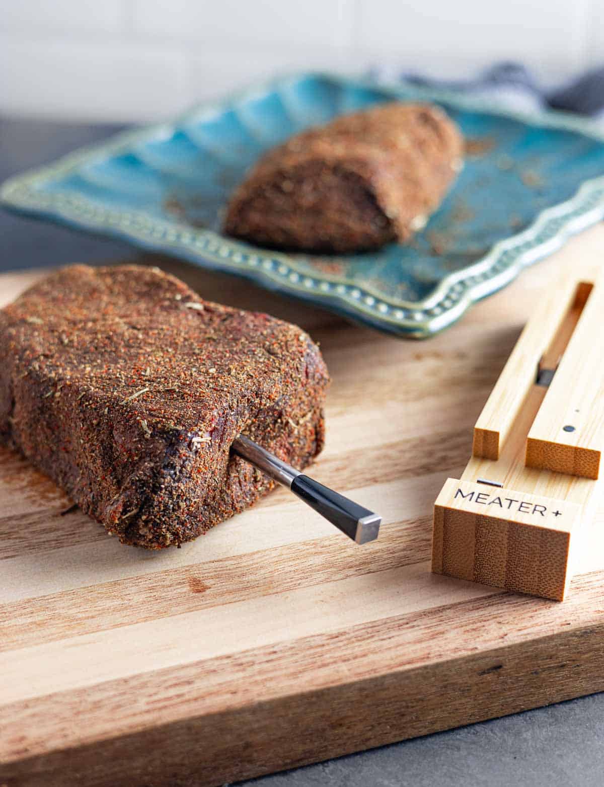 Raw rubbed sirloin steak with a Meater smart thermometer inserted in one end with the case and a steak on a plate behind.