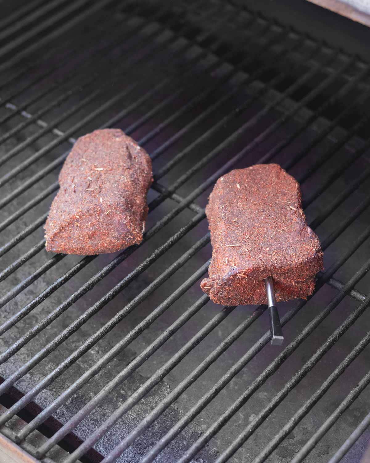 Two raw dry-rubbed sirloin steaks on the smoker grates ready to smoke.