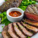 Partially sliced sirloin steak on a plate with cilantro garnish and small white bowl of red chimichurri sauce.