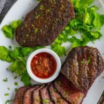 Smoked sirloin steaks sliced on a platter with ramekin of chimichurri sauce.