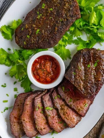 Smoked sirloin steaks on a white platter with slices, red chimichurri sauce and a cilantro garnish.