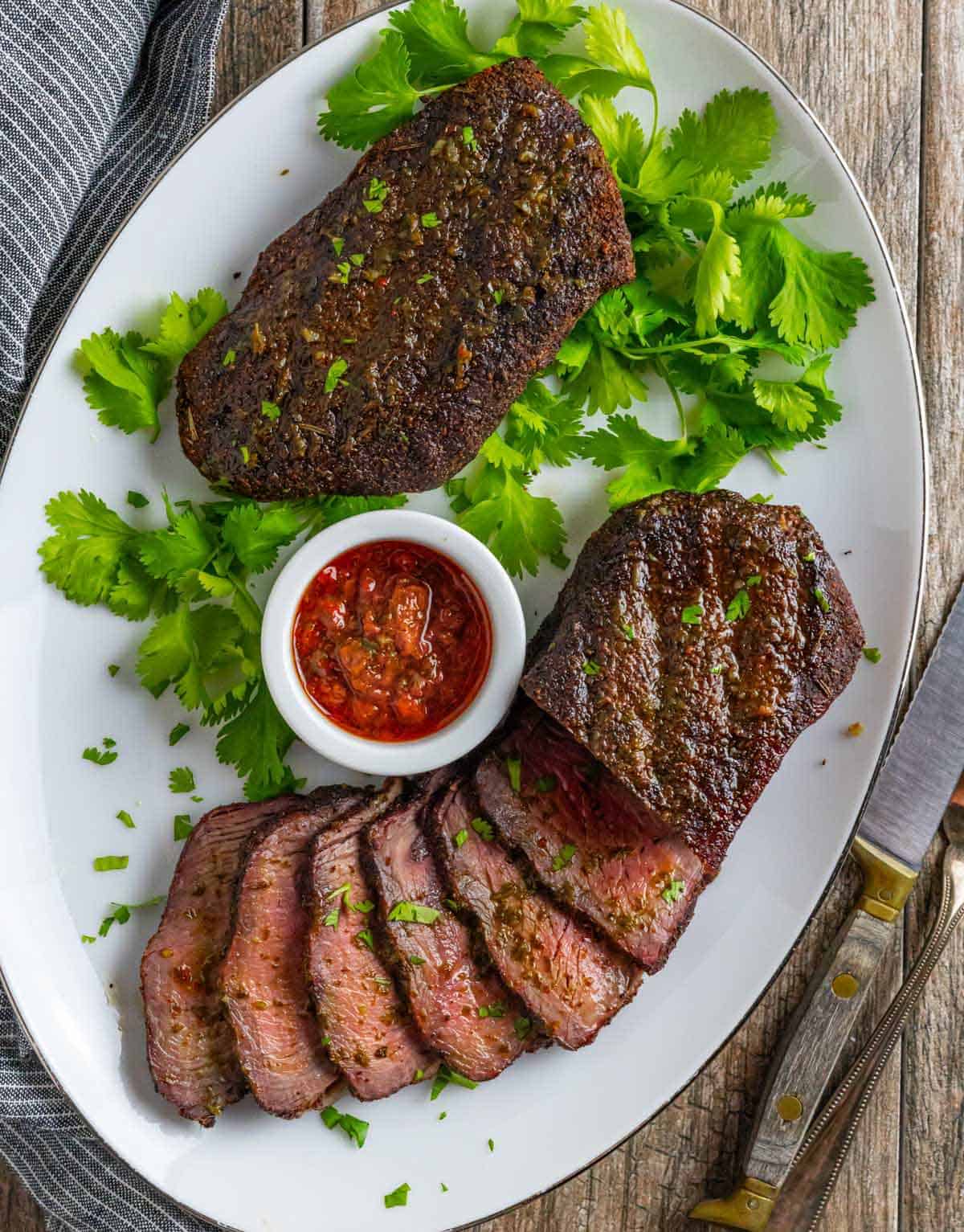 Smoked sirloin steaks sliced on a platter with ramekin of chimichurri sauce.