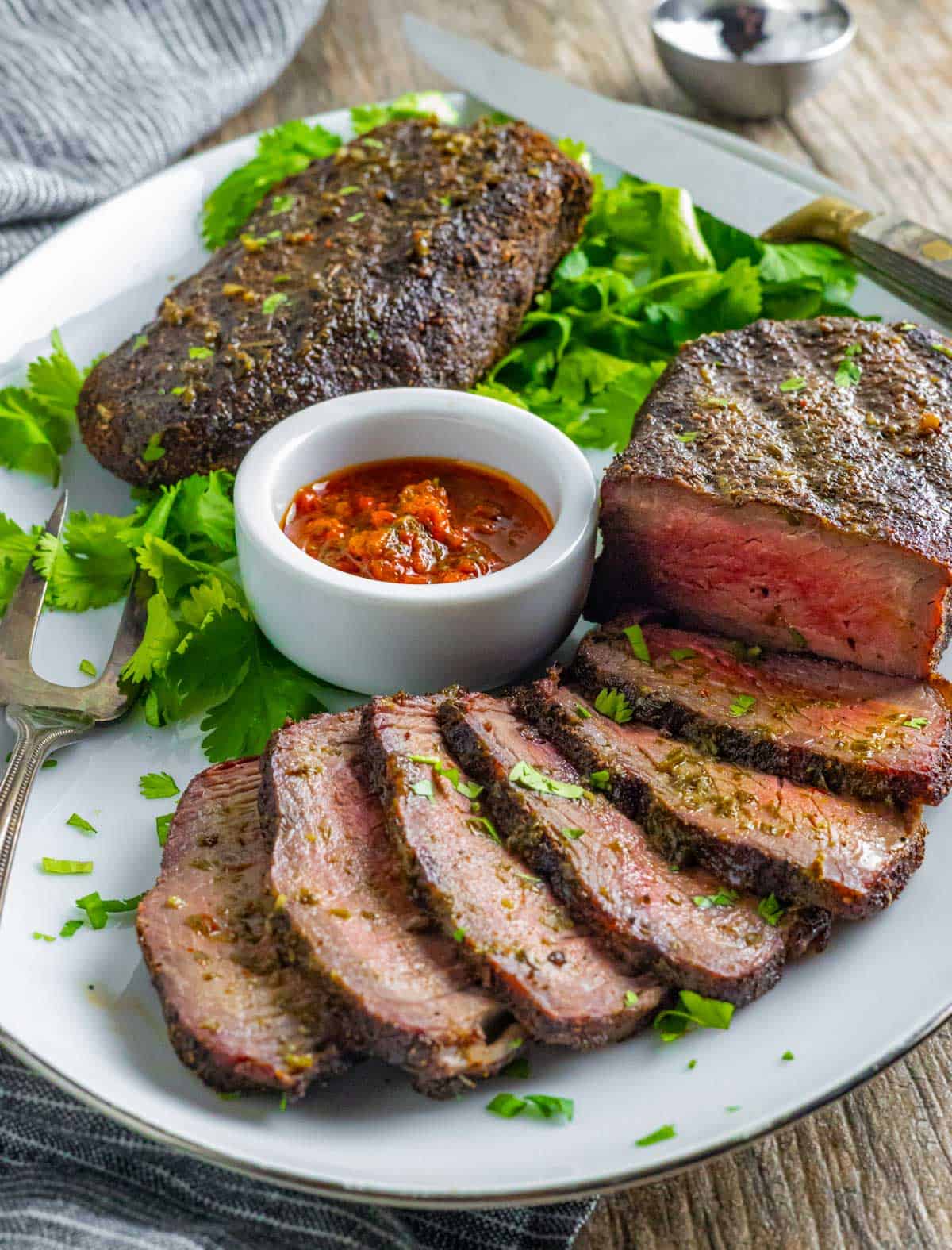 Partially sliced sirloin steak on a plate with cilantro garnish and small white bowl of red chimichurri sauce.