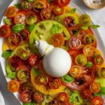 Oval plate of caprese including sliced cherry and multi colored tomatoes, a ball of burrata garnished with fresh basil.