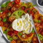 Burrata caprese salad on a large oval plate with burrata ball cut open and drizzled with olive oil.