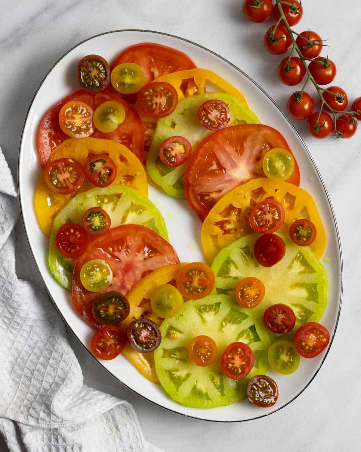 Oval plate with sliced cherry tomatoes and large tomatoes of multiple colors layered over each other.
