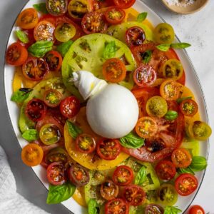 Plate of burrata caprese salad of multi-colored sliced large and cherry tomatoes with a burrata cheese ball in the center.