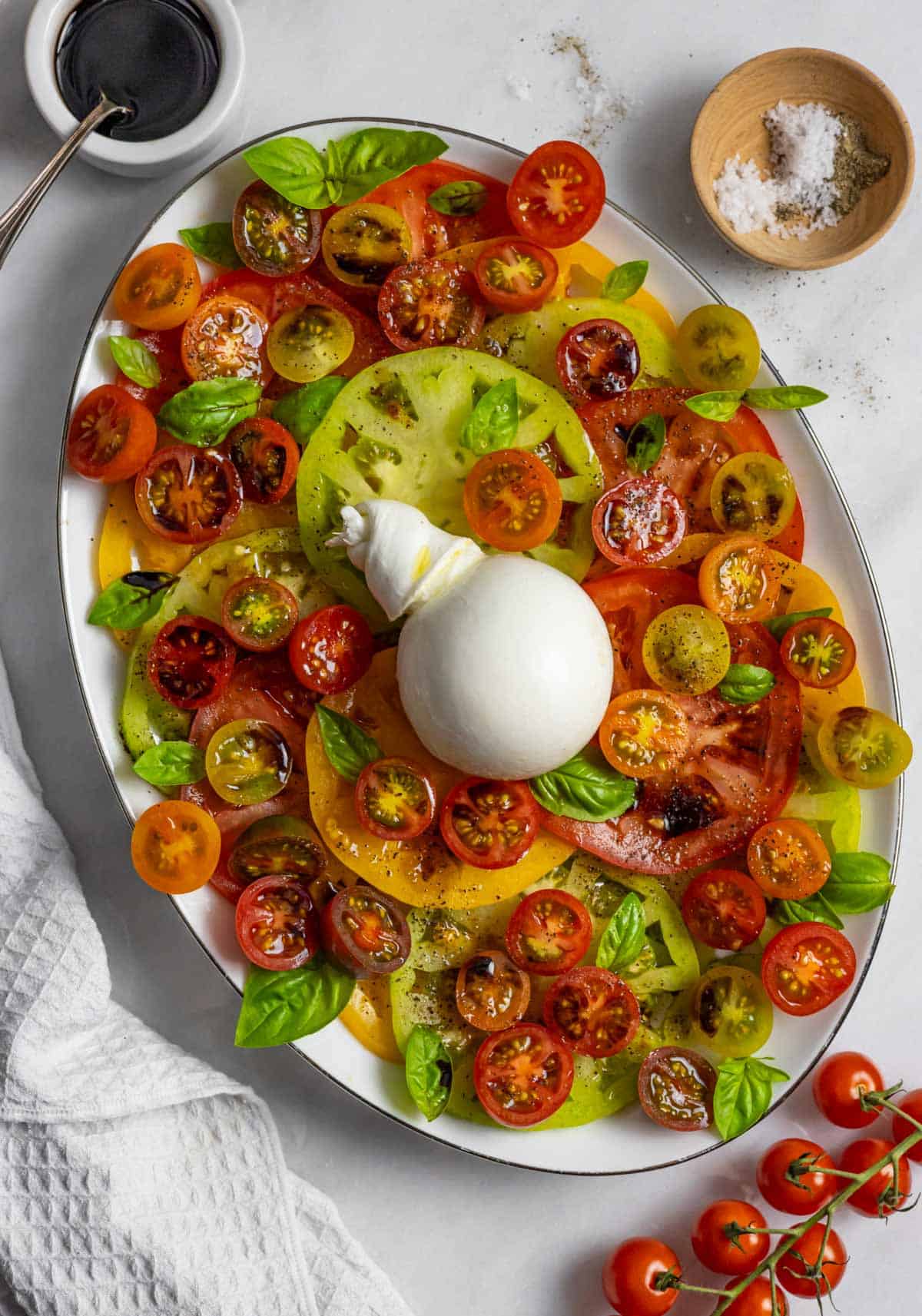 Oval plate of caprese including sliced cherry and multi colored tomatoes, a ball of burrata garnished with fresh basil.