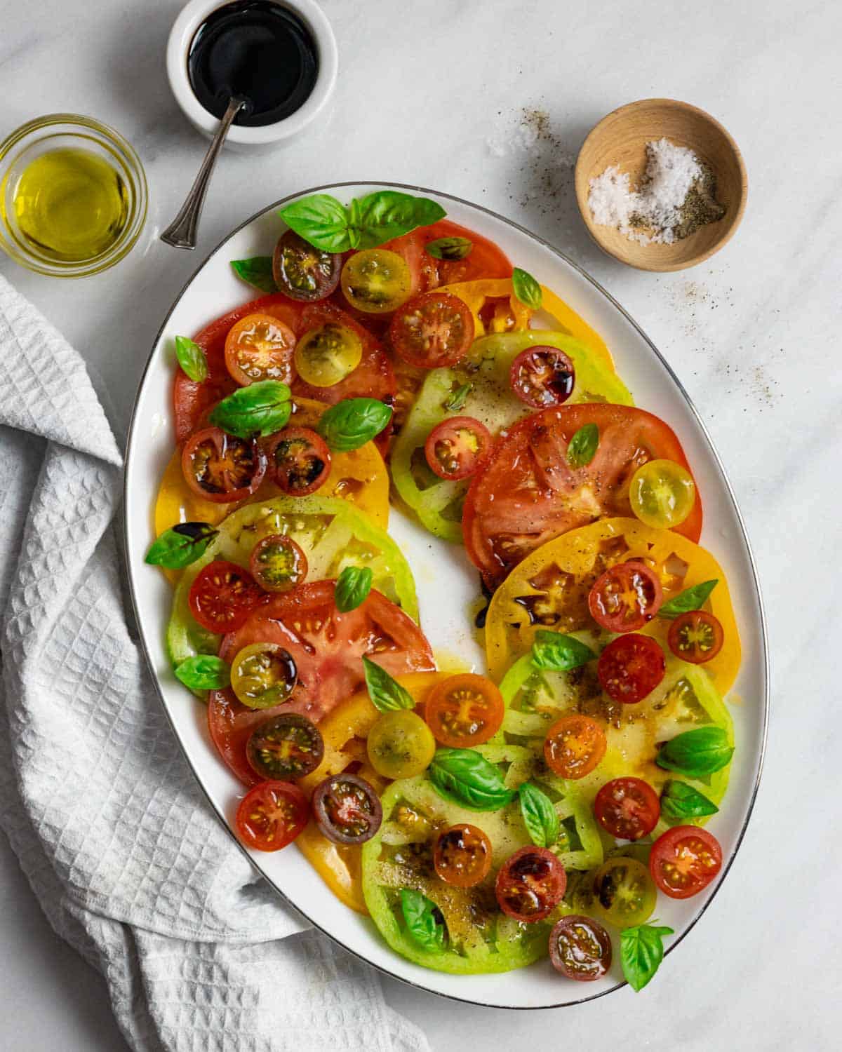 Plated caprese including sliced colorful tomatoes, balsamic reduction, seasoning and fresh basil leaves.