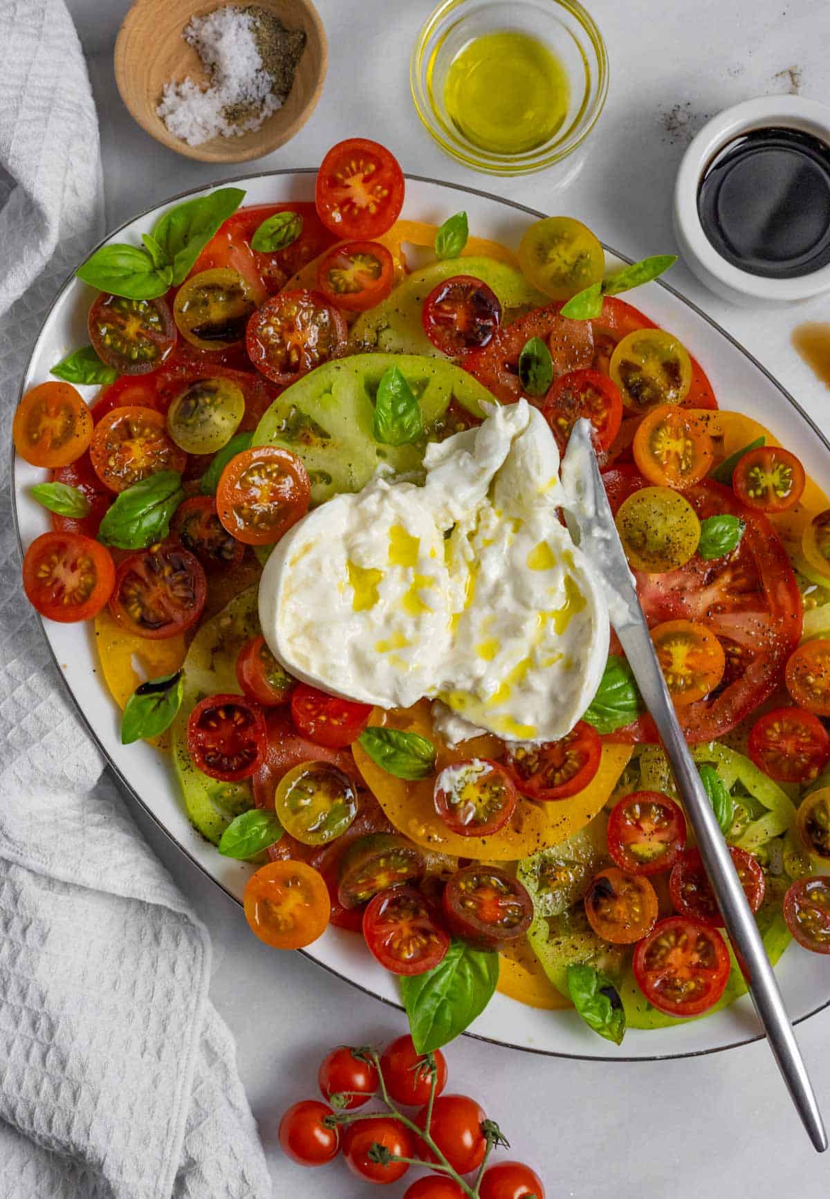 Burrata caprese salad on a large oval plate with burrata ball cut open and drizzled with olive oil.