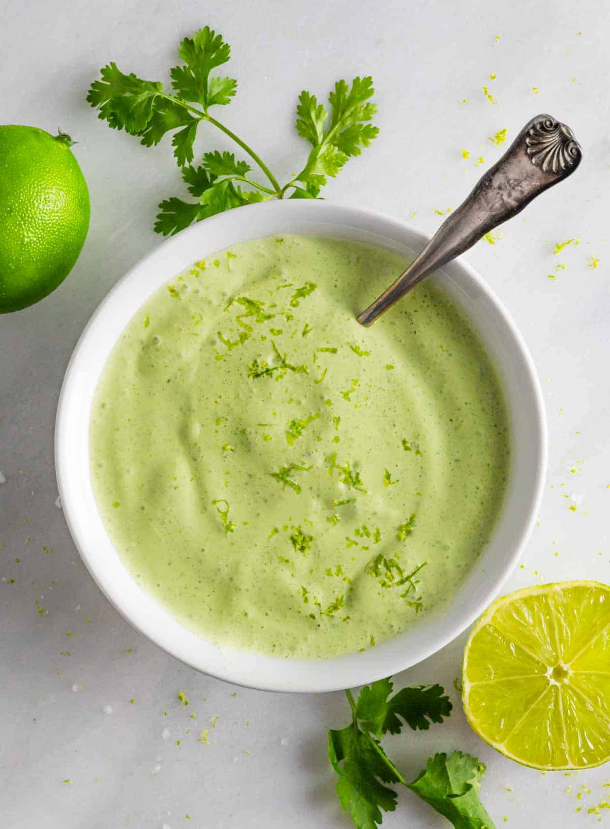 Blended cilantro lime crema in a white bowl with a spoon with half and whole lime next to it.