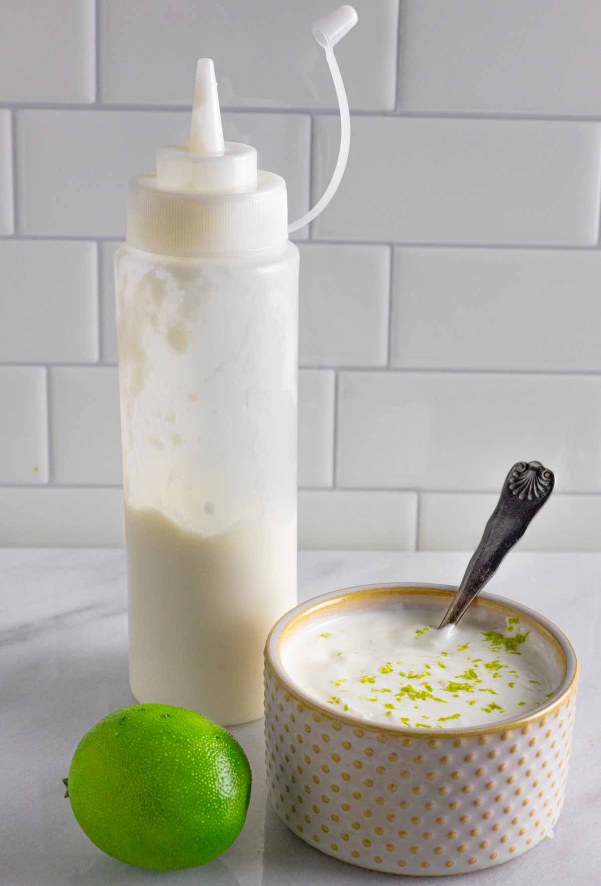 Lime crema in a bowl with lime zest on top and in a squeeze bottle of sauce on a marble board.