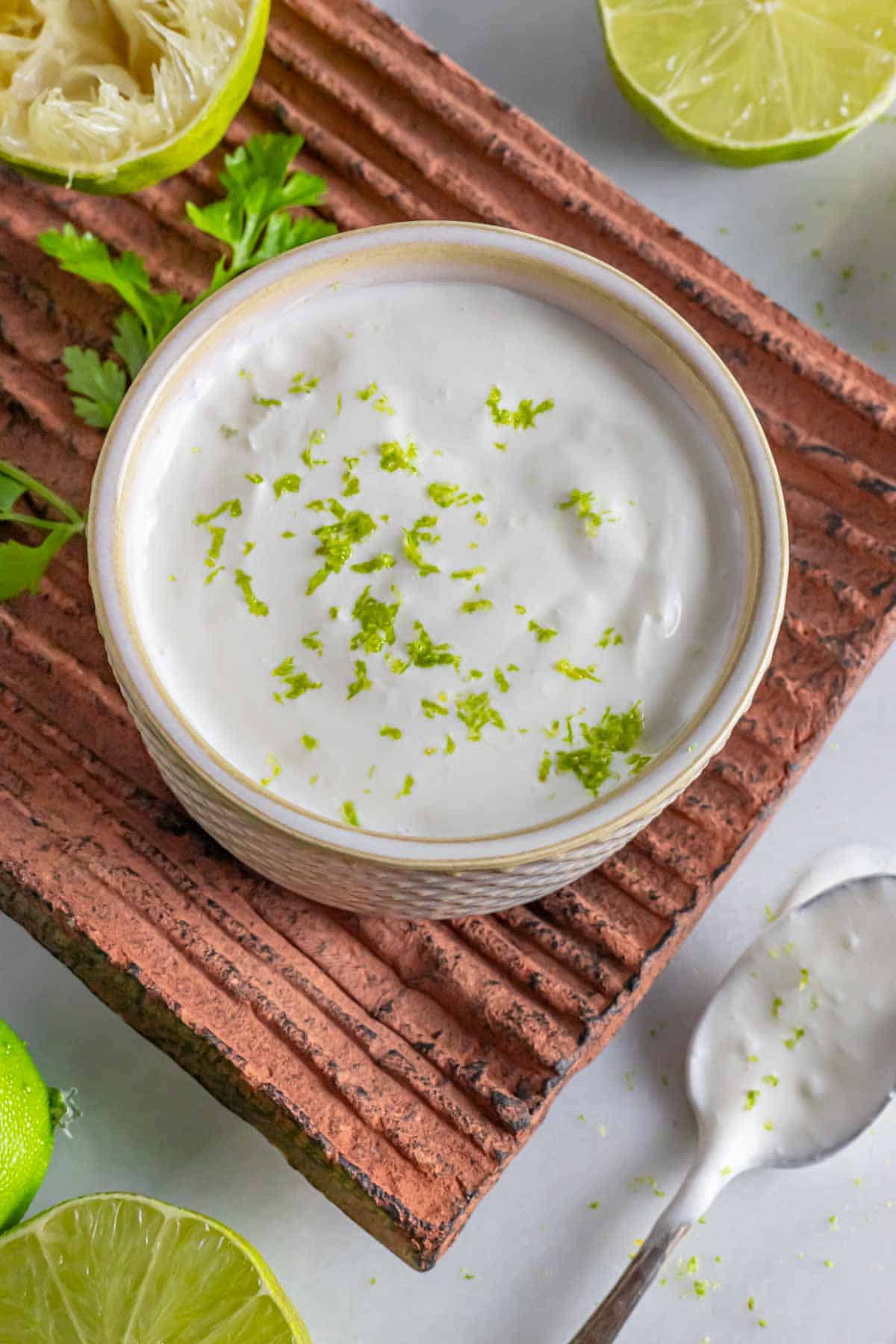 White bowl of crema topped with lime zest on a clay platter with lime garnish and sauce covered spoon.