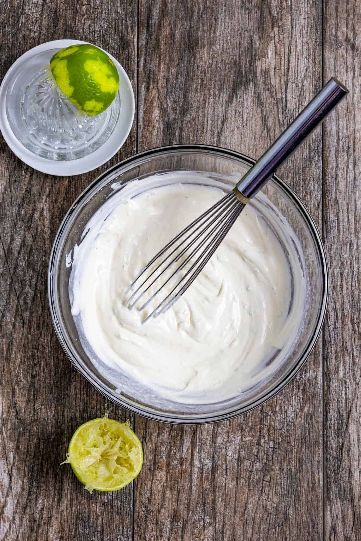 Lime crema mixed in a glass bowl with a whisk in it and squeezed lime next to the bowl.